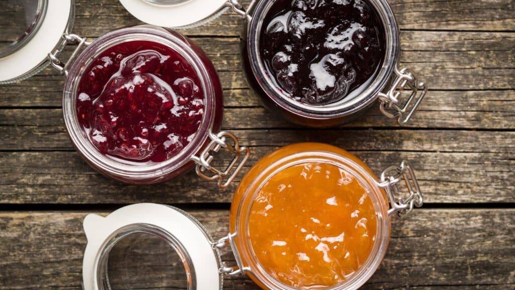 Assorted fruit preserves in open jars on wooden surface.