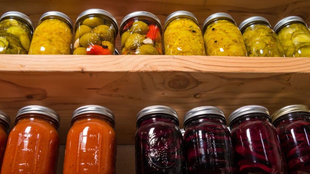 Assorted pickled vegetables in jars on wooden shelf.